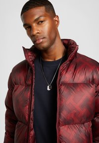 Pufjakke i mørkerød med et mønstret tekstur, lynlåslukning foran og høj krave, båret over en sort skjorte. Halskæde synligt på modellen.