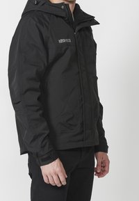 Svart jacka med dragkedja framtill, justerbar huva, sidofickor och kardborreärmar. Tillverkad av ett glansigt, lätt material.