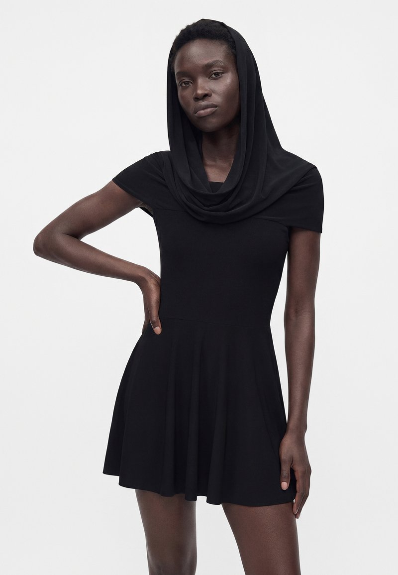Femme portant une courte robe noire à encolure à capuche, debout, une main sur la hanche, sur un fond clair uni.