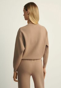 Beżowy sweter robiony na drutach z wysokim dekoltem, opadającymi ramionami i luźnym krojem, w zestawieniu z pasującymi spodniami. Gładka tekstura, minimalistyczny design.