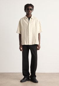 Linnen beige overhemd met korte mouwen en knopen, gecombineerd met zwarte broek en zwarte sneakers. Model draagt een donkere zonnebril en staat tegen een lichte achtergrond.