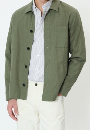 Homme portant une veste boutonnée vert olive sur une chemise blanche et bleue à rayures, associée à un pantalon blanc, main dans la poche.