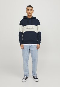 Sudadera con capucha en azul marino y crema, con un bolsillo frontal y logo bordado. Combinada con jeans de color azul claro y zapatillas negras, de pie frente a un fondo simple.