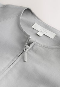 Vêtement en lin gris clair avec encolure ronde et fermeture éclair argentée partiellement ouverte au centre devant.