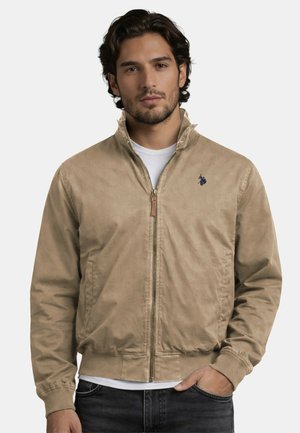 Man wearing a beige zip-up jacket over a white shirt, standing with one hand in pocket against a plain background.