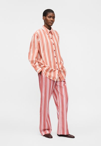Γυναίκα στέκεται με τα χέρια στις τσέπες, φοράει μια φαρδιά πορτοκαλί και λευκή κάθετα ριγέ πουκάμισο και ταιριαστό ριγέ παντελόνι με καφέ παπούτσια.