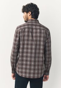 Chemise boutonnée à carreaux marron avec manches longues, dotée d'un col classique, d'un empiècement au dos et de poignets boutonnés. Le tissu présente une finition texturée.