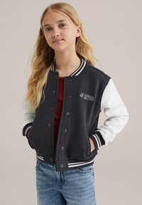 Črno-bela univerzitetna jakna s pritisnimi gumbi, črtastimi manšetami in napisom "AU REVOIR" na prsih. Mehka tkanina, sproščen kroj.