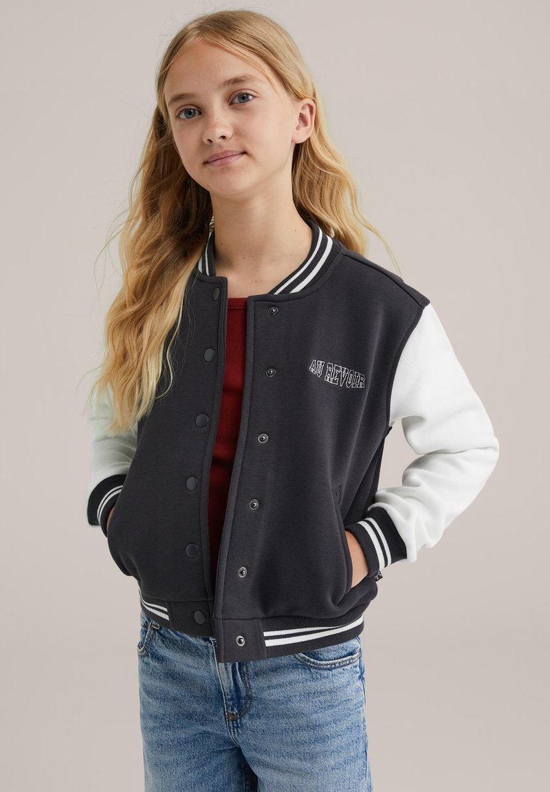 Črno-bela univerzitetna jakna s pritisnimi gumbi, črtastimi manšetami in napisom "AU REVOIR" na prsih. Mehka tkanina, sproščen kroj.