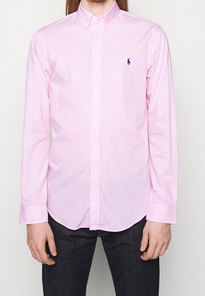 Mężczyzna w różowo-białej koszuli na guziki z pionowymi paskami i kołnierzykiem oraz ciemnych spodniach, ręce swobodnie opuszczone wzdłuż ciała.