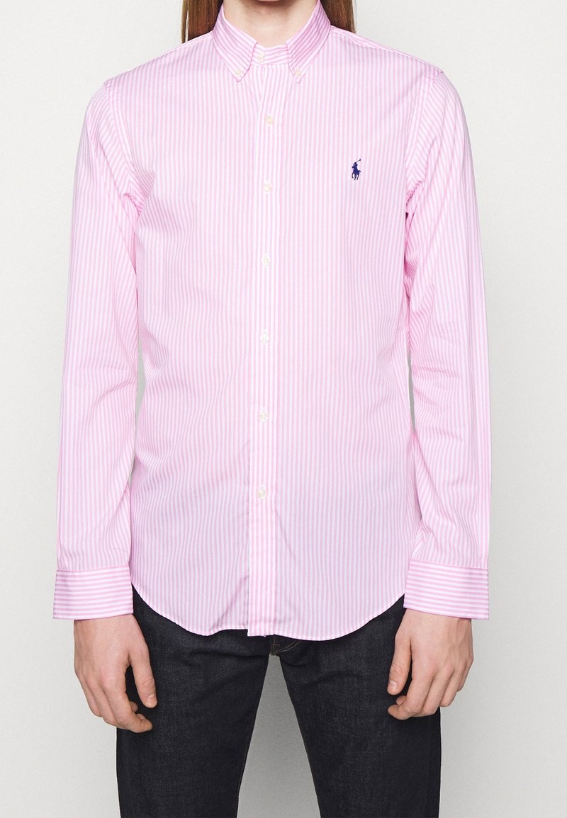 Mann trägt ein rosa-weiß gestreiftes Hemd mit Kragen und dunkle Hose, die Hände entspannt an den Seiten.