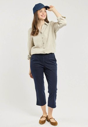 Femme portant une chemise beige boutonnée, un pantalon court bleu marine, des ballerines marron et une casquette bleu marine, souriante et ajustant sa casquette d'une main.