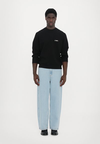 Sweatshirt noir avec logo, associé à un jean large bleu clair et des chaussures noires. Le mannequin se tient devant un fond blanc uni.