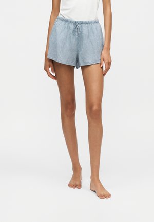 Kvinde iført hvid top og løstsiddende lyseblå ternede shorts med snøre, stående barfodet på en ensfarvet hvid baggrund.