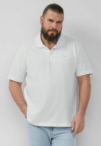 Weißes Polo-Shirt aus Baumwolle mit klassischem Kragen, kurzen Ärmeln und einem dezenten Logo auf der Brust. Kombiniert mit hellblauen Jeans.