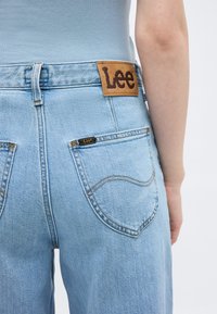 Πίσω όψη ατόμου που φοράει ανοιχτό μπλε τζιν Lee με ψηλή μέση, με ετικέτα της μάρκας στη ζώνη και στην πίσω τσέπη, ανοιχτό μπλε τοπ, απλό φόντο.