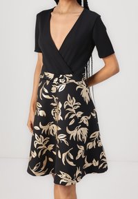 Svart klänning med djup V-ringning och korta ärmar, med en flödande kjol prydd med guldbladsmönster. Slät textur och passform.