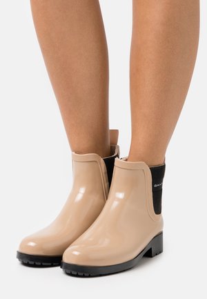 Bottes cheville en caoutchouc beige à finition brillante, avec des panneaux latéraux élastiques noirs et une semelle noire robuste. Dotées d'une linguette à l'arrière.