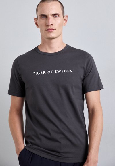 Tamsiai pilka medvilnės marškinėliai su trumpomis rankovėmis, su balta tekstu "TIGER OF SWEDEN" centriniu būdu ant priekinės dalies. Sklandi tekstūra.