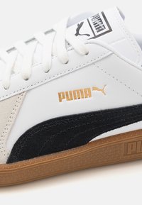 Zapatilla blanca con materiales de ante y piel; presenta acentos negros, un logotipo dorado y suela de goma texturizada. Cierre de cordones en la parte superior.