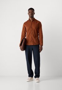 Cămașă maro cu nasturi, cu mâneci lungi, pantaloni negri închis, adidași bej și o geantă maro. Textură de țesătură netedă, design simplu.