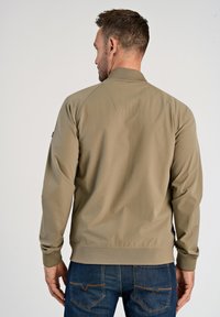 Giacca khaki con zip, colletto a coste, polsini elasticizzati e superficie testurizzata. Mostrata dal retro, abbinata a jeans blu scuro.