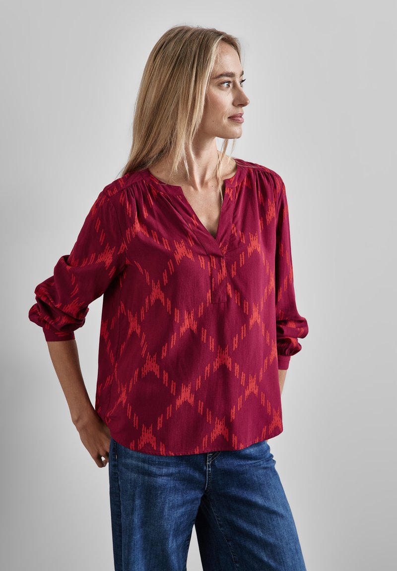 Blusa rossa a motivo con scollatura a V, dettagli arricciati sulle spalle e maniche lunghe a sbuffo, realizzata in tessuto leggero. Indossata con jeans blu.