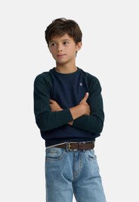 Sudadera azul marino con mangas verde oscuro, puños acanalados y un pequeño logo en el pecho, combinada con jeans azul claro y un cinturón marrón.