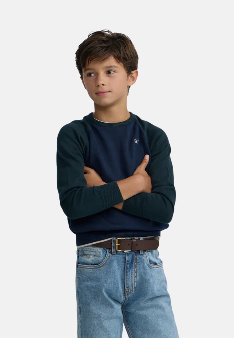Sudadera azul marino con mangas verde oscuro, puños acanalados y un pequeño logo en el pecho, combinada con jeans azul claro y un cinturón marrón.