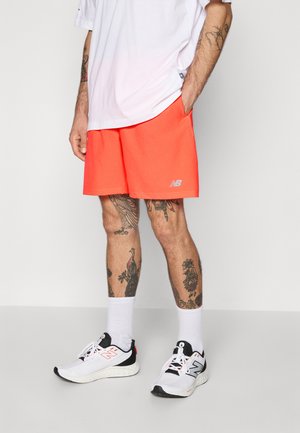 Mann trägt leuchtend orangefarbene Sportshorts, weiße New Balance Sneakers, weiße Socken und ein weißes Shirt, steht mit den Händen in den Taschen.