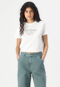 Hvit bomulls t-skjorte med korte ermer, utsmykket med et rhinestones-beriket "Carhartt WIP"-logo. Kombinert med høyhalsede grønne bukser.