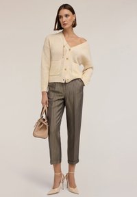 Pletený kardigan v krémové barvě s otevřeným pletením a dvěma předními kapsami, spárovaný s olivově zelenými pruhovanými kalhotami a béžovými lodičkami na vysokém podpatku.