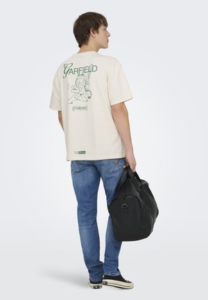 Joven con camiseta beige de Garfield, vaqueros azules, zapatillas negras, sosteniendo una bolsa de deporte negra, de pie frente a fondo blanco.