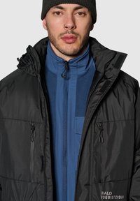 Czarna ocieplana kurtka z kapturem, z dwoma kieszeniami na zamek, noszona na niebieskim polarze z suwakiem i kieszenią na klatce piersiowej.