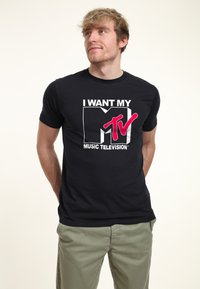 Black cotton T-shirt featuring "I WANT MY MTV" in white and pink text, crew neckline, short sleeves, and a relaxed fit.
