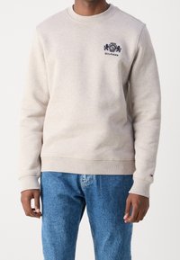 Light beige sweatshirt made of soft fabric, featuring a round neck, ribbed cuffs, and a navy embroidered logo on the chest. Denim jeans visible below.