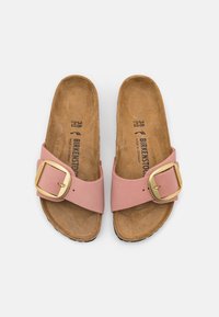 Sandales Birkenstock roses avec une large sangle en cuir, dotées d'une grande boucle en métal doré et d'une semelle en daim sur une semelle en caoutchouc marron.