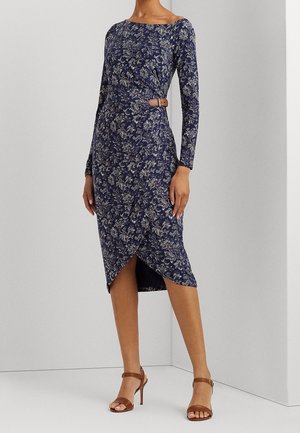 Vrouw die een marineblauwe midi-jurk met lange mouwen en bloemenprint draagt, met een bruine taille riem en bruine sandalen met hoge hakken, staand tegen een effen muur.