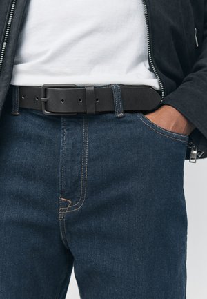 Personne portant un jean bleu foncé avec des surpiqûres orange, une ceinture en cuir noire avec une boucle mate, une chemise blanche et une manche de veste noire visible.