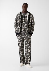 Camouflage trainingspak in grijs en zwart met een capuchon en lange broek. Het materiaal lijkt zacht, met contrasterende zwarte accenten en witte logo's.