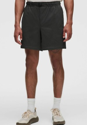 Mand iført sorte elastiske shorts med bælte, hvide sokker og beige træningssko, der står mod en neutral baggrund.