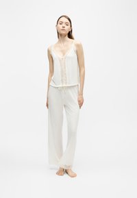 Jeune femme debout pieds nus portant un caraco blanc orné de dentelle et un pantalon de pyjama ample sur un fond clair uni.