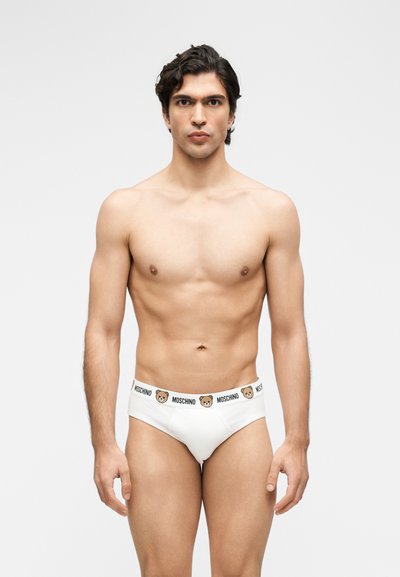Νέος άνδρας με σκούρα μαλλιά, όρθιος χωρίς πουκάμισο, φορώντας λευκά εσώρουχα Moschino με λογότυπο αρκουδάκι στη λαστιχένια μέση.