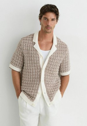 Man wearing a brown and white patterned short-sleeve knit cardigan over a white tank top and white pants, standing with hands in pockets.