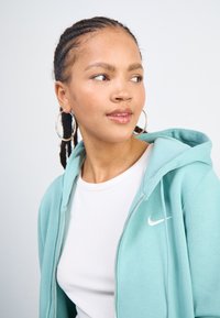Nike zip-up hoody gaišā tirkīza krāsā ar baltu logotipu, kopā ar baltu ribotu T-kreklu, valkāts ar lieliem sudraba apli auskariem.