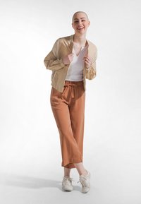 Giacca bomber beige sopra una maglietta bianca a scollo a V, abbinata a pantaloni a gamba larga color ruggine e sneakers bianche. Tessuto liscio, vestibilità rilassata.