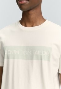Camiseta de manga corta y cuello redondo en algodón beige claro con un gráfico en verde menta que dice "DENIM TOM TAILOR" y un fondo texturizado.