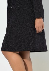 Schwarzes Kleid aus glitzerndem, strukturiertem Stoff, mit langen Ärmeln und einem leicht ausgestellt geschnittenen Saum. Sichtbare Hand, die den Stoff an der Taille hält.