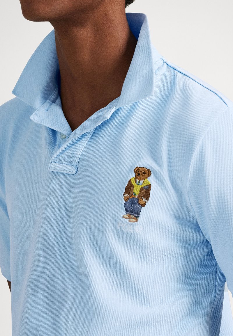 Nahaufnahme einer Person, die ein hellblaues Poloshirt mit hochgestelltem Kragen trägt, auf der Brust ist ein gestickter Bär in Freizeitkleidung zu sehen.