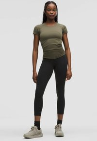 Top fitted a maniche corte di colore verde oliva con una finitura testurizzata, abbinato a leggings neri. La modella indossa sneakers grigio chiaro e calzini scuri.